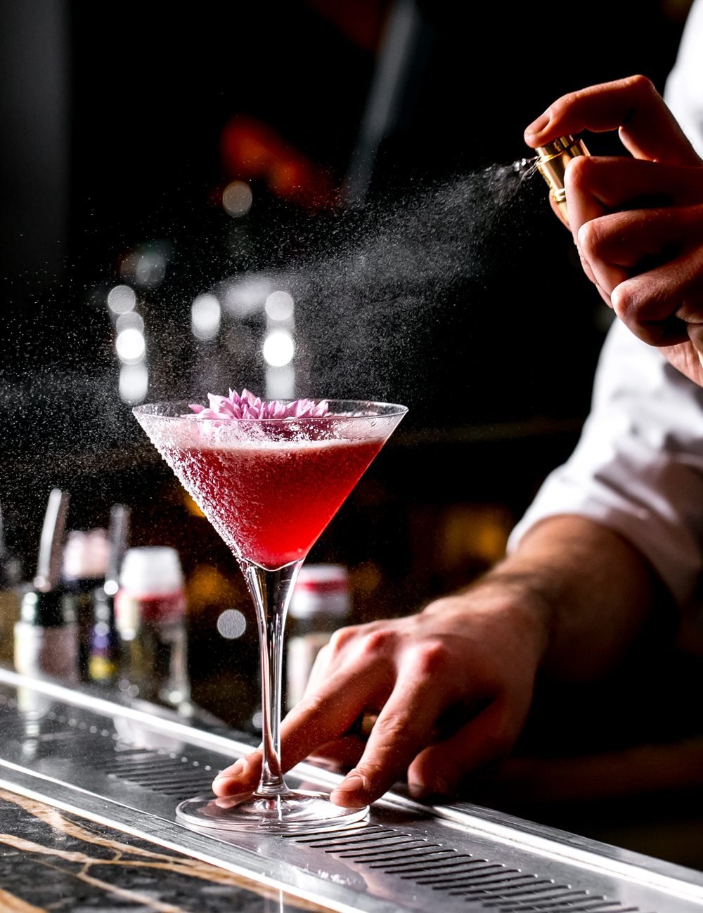 Bartender rociando un aroma con atomizador dorado sobre una copa con flor comestible